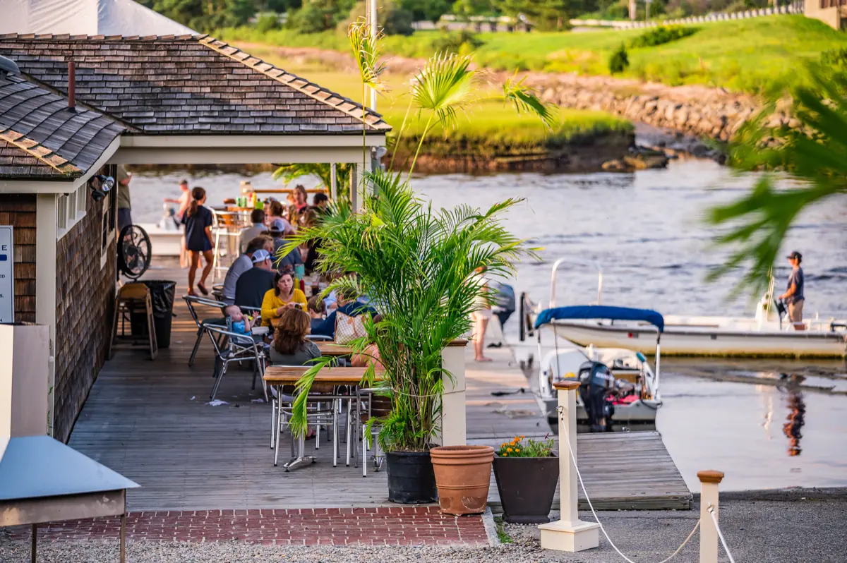 Guests dining on the waterfront deck with boats moored beyond in evening light