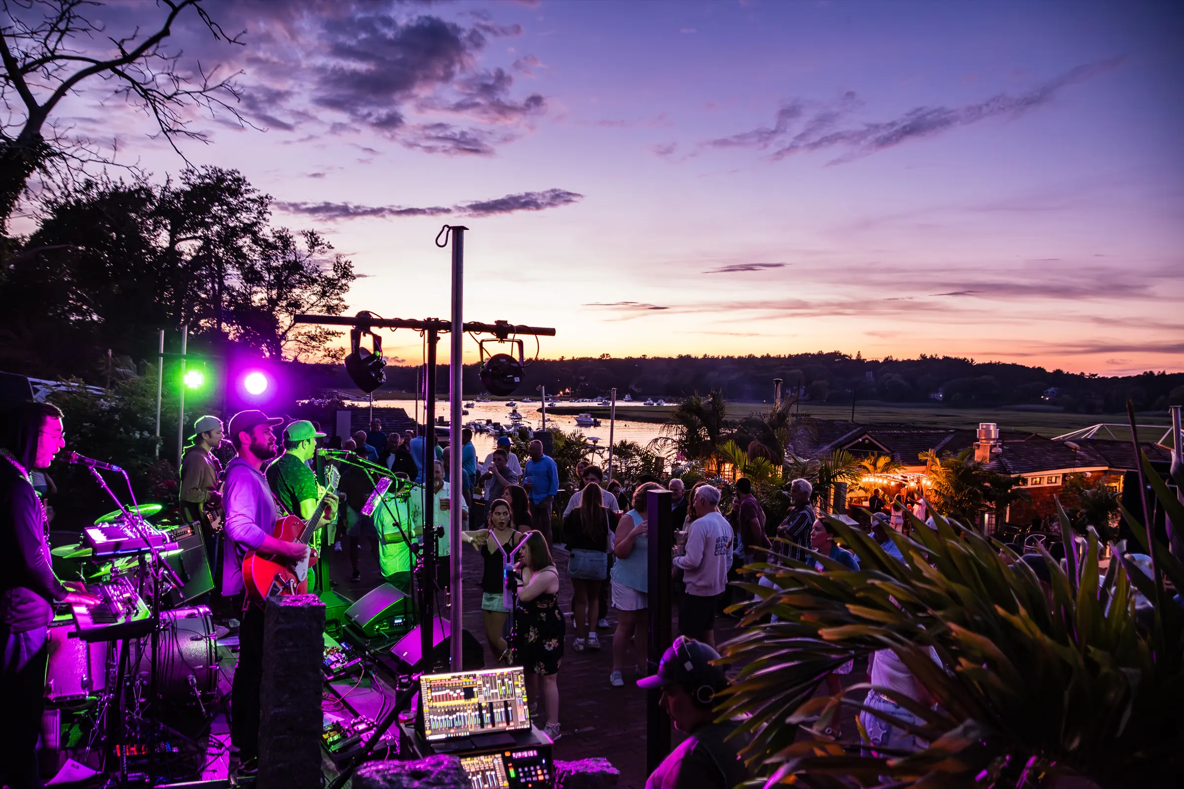 A band playing to a crowd on the waterfront under a vivid purple sunset
