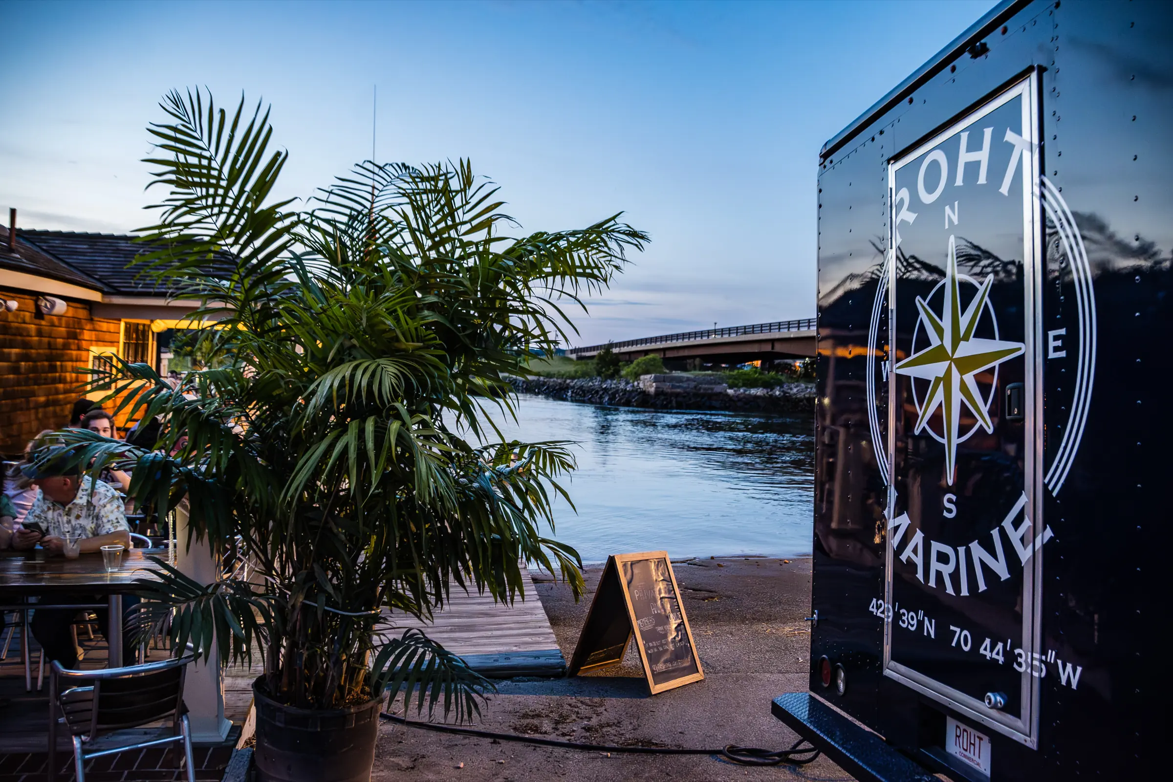The ROHT Marine trailer bar beside a palm and the Green Harbor bridge at dusk