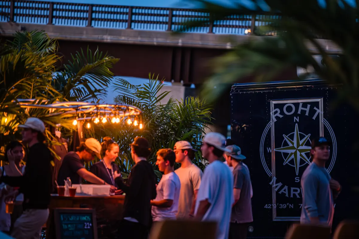 A crowd gathered at the trailer bar under string lights with the harbor bridge behind